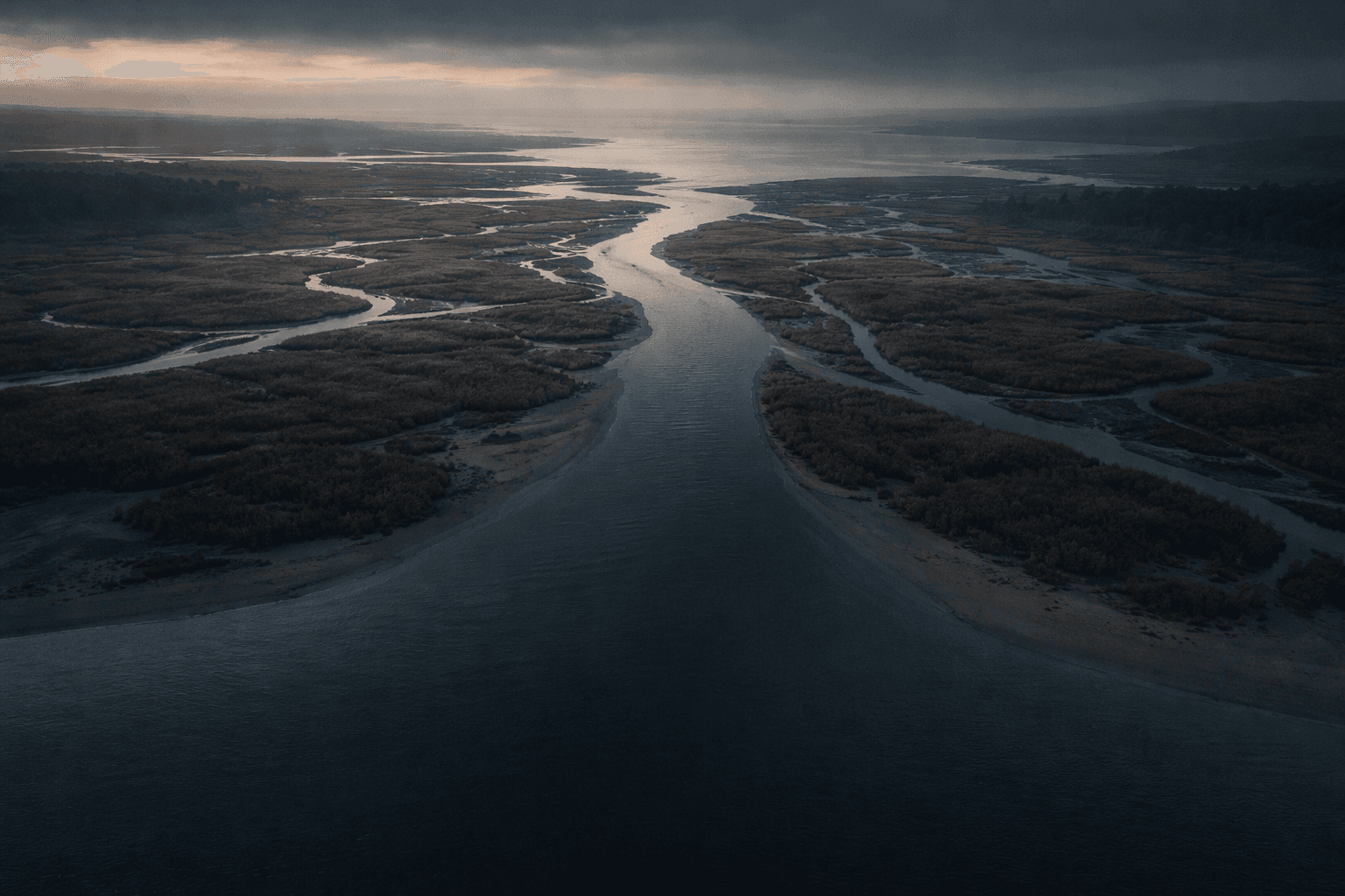 Aerial dusk view of a braided river funneling through muted marshes, with an empty sandy shoreline occupying the quiet lower third.