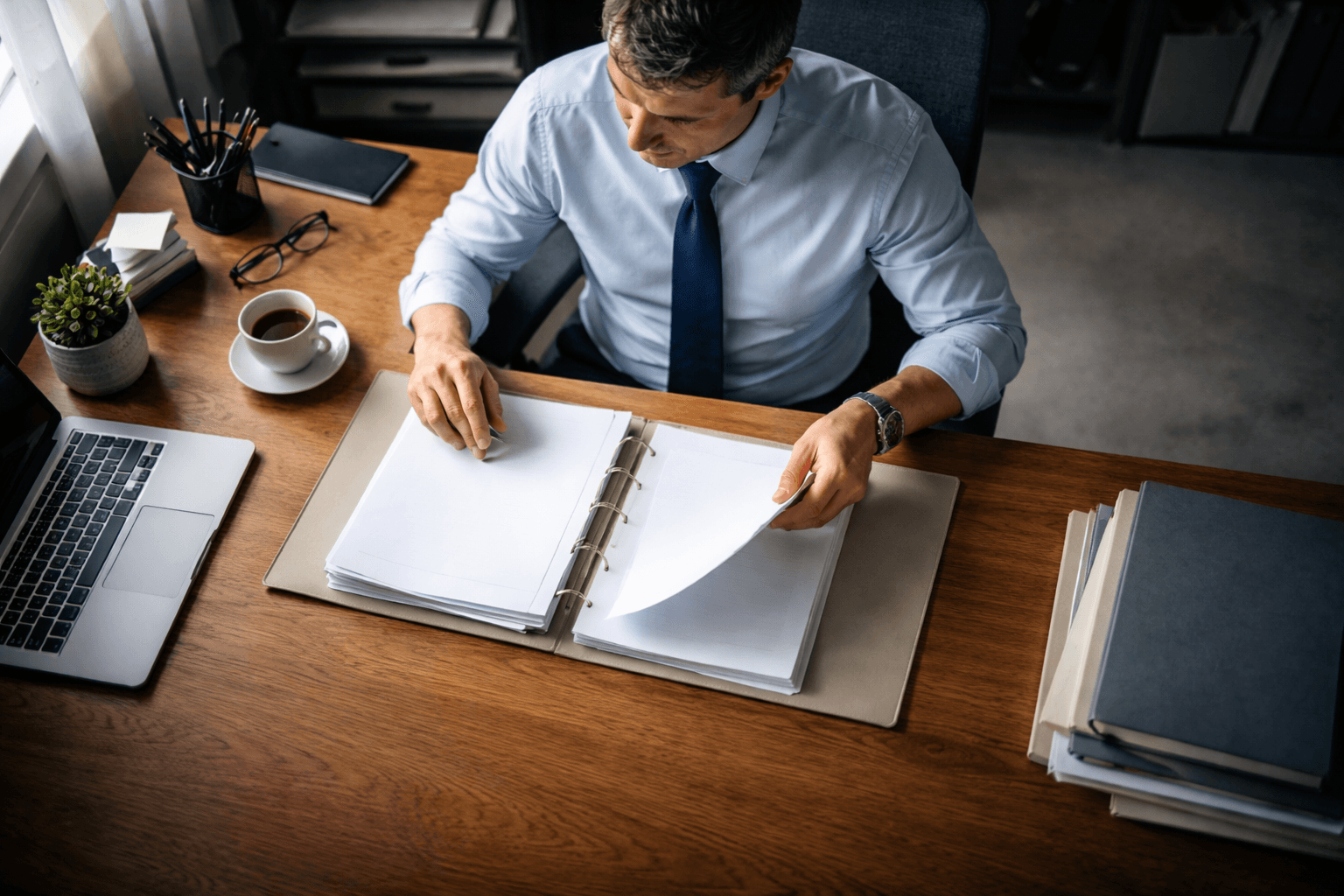 High-angle view from above of a fictional compliance officer at a wooden desk, sorting blank papers and a neutral binder under soft morning light; the person's face is visible but anonymous, with the upper two-thirds filled and the lower third calm and uncluttered.